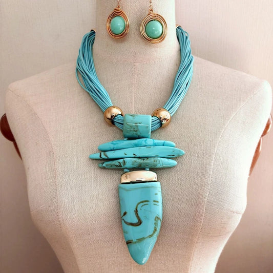 Turquoise statement necklace with a large teardrop pendant, layered cord detail, and gold-tone accents, displayed on a beige fabric mannequin torso with matching drop earrings visible at the top.