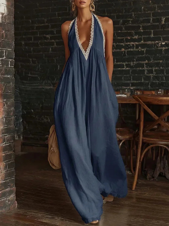 A woman wearing a flowing navy blue halter maxi dress with a deep V-neckline and white lace trim, standing in a rustic room with a dark brick wall and wooden furniture.