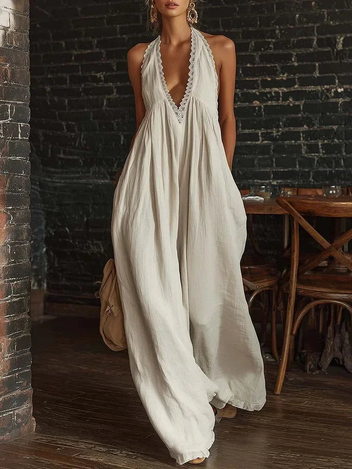 A woman models a sleeveless, off-white maxi dress with a deep V-neckline and scalloped trim, set against a rustic dark brick wall and wooden table background.