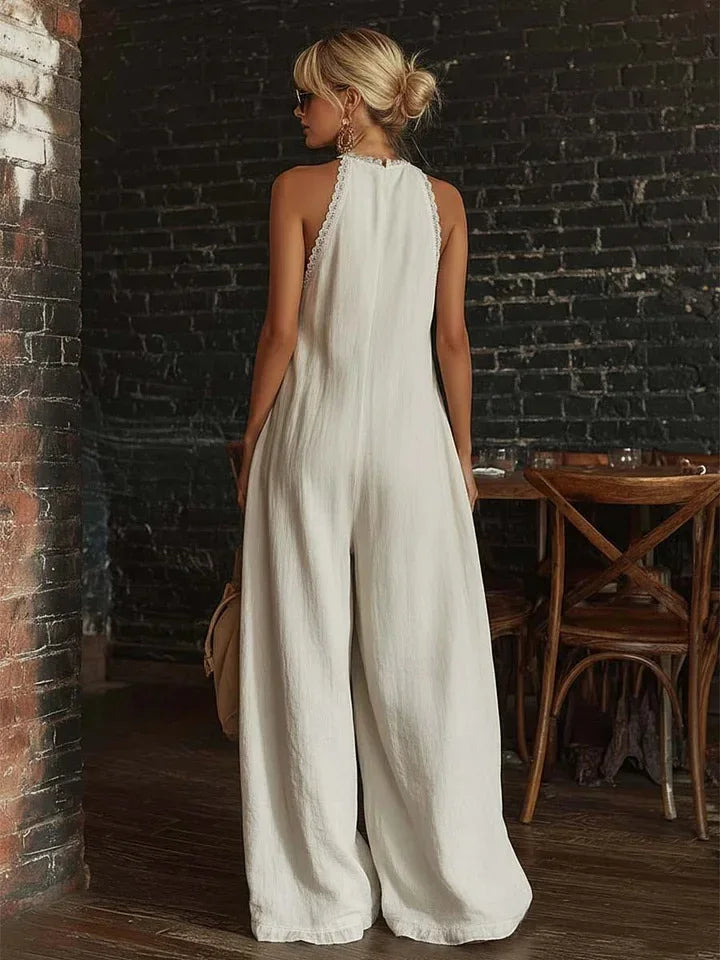 Woman in a white jumpsuit standing in a rustic indoor setting with brick walls and wooden furniture.