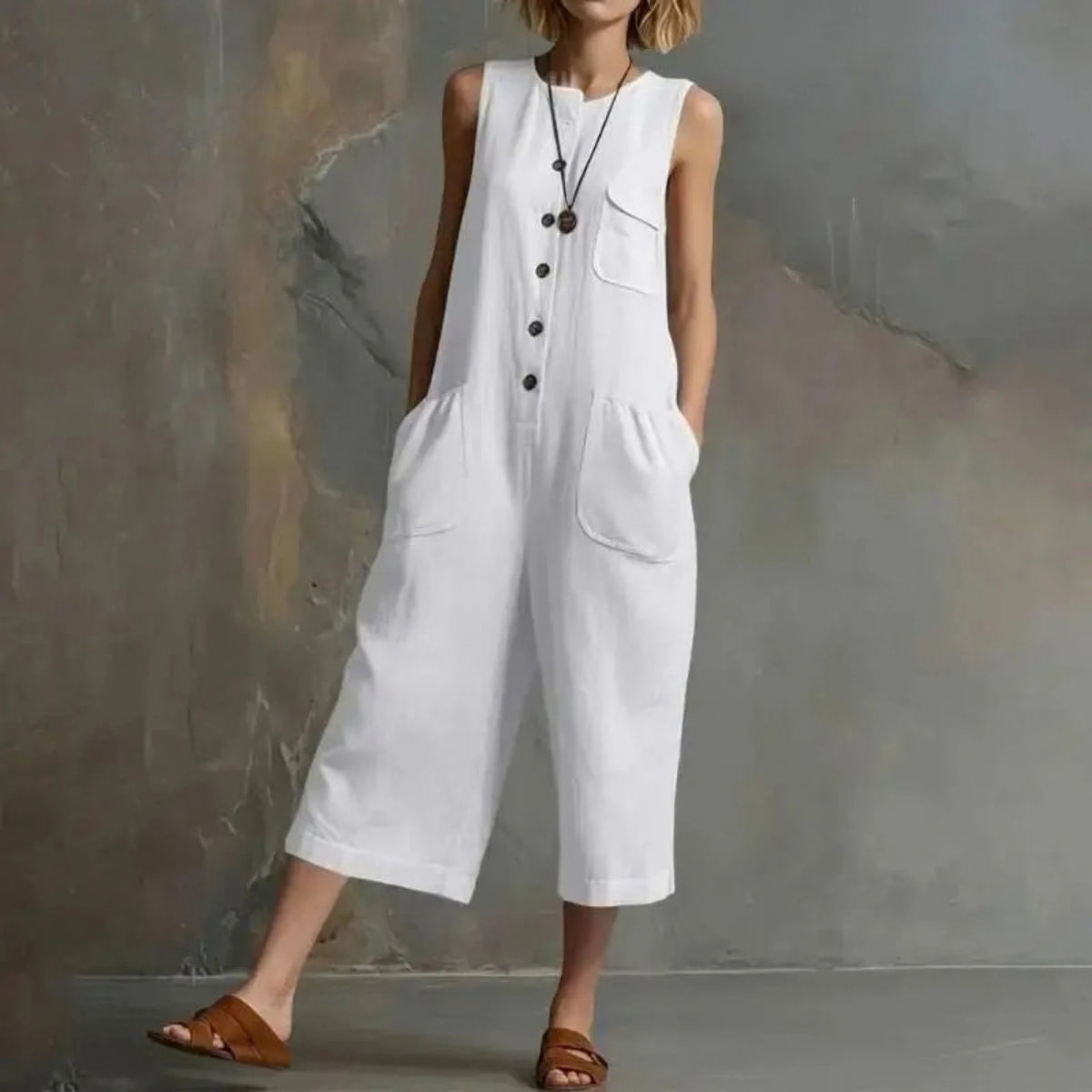 A woman wearing a white sleeveless linen-look jumpsuit with front buttons, chest pocket, and large patch pockets, standing against a textured gray wall. The brand name XJGOOD is printed in black in the upper left corner.
