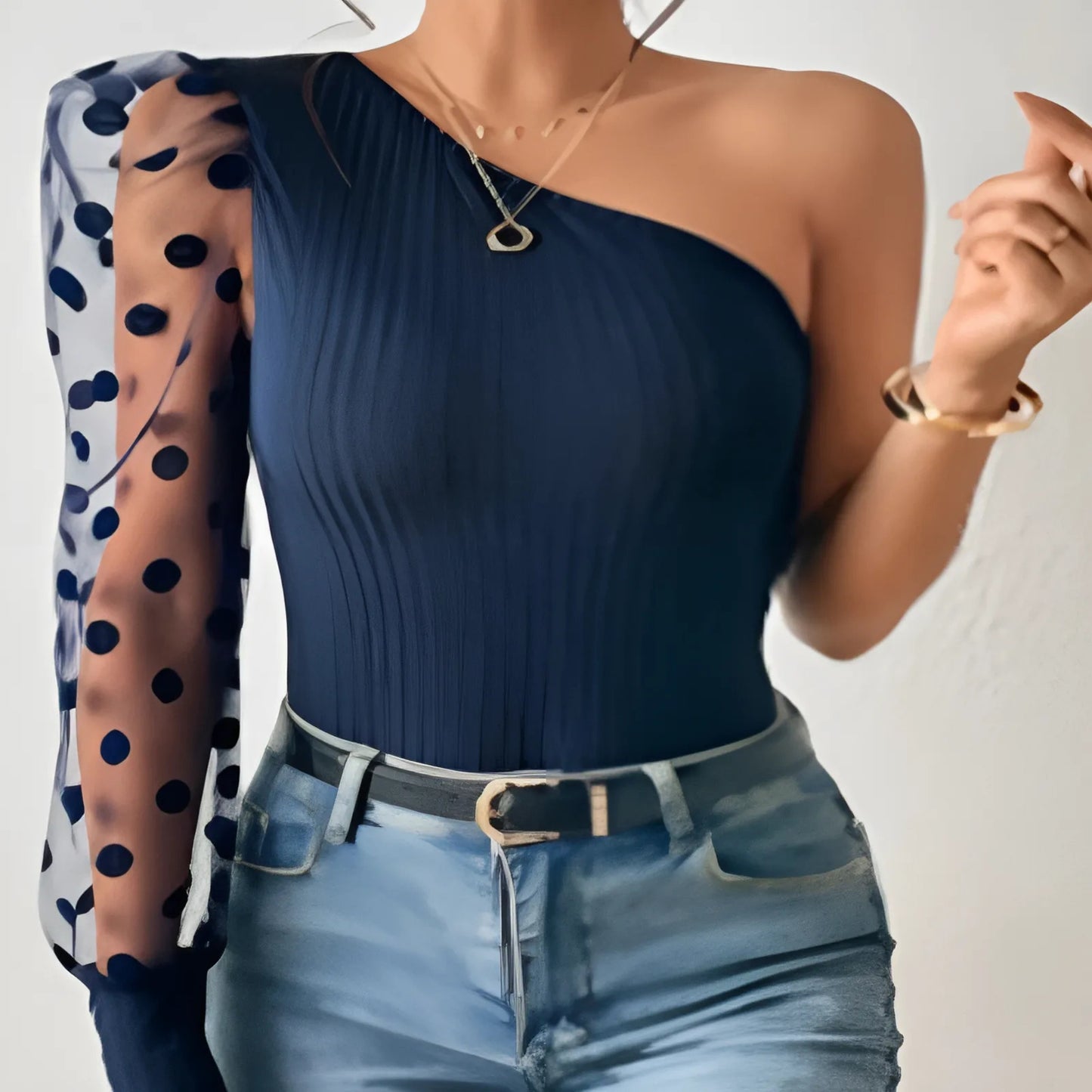 Woman wearing a navy one-shoulder top and blue jeans with a sheer polka dot sleeve.
