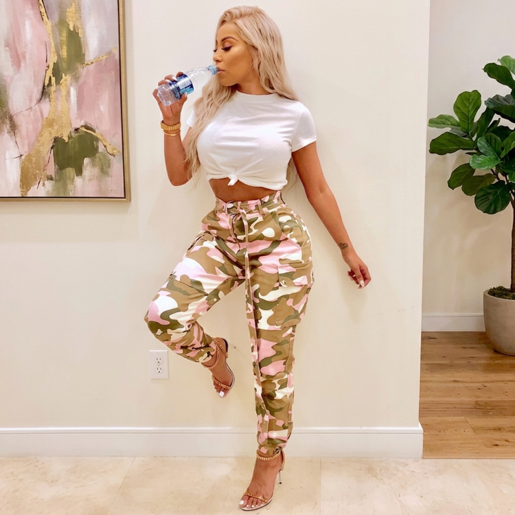Women a camouflage pant standing next to a wall sipping on a bottle of water.