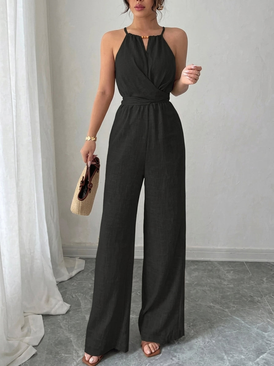 A woman modeling a black sleeveless halter jumpsuit with a wrap-style bodice and wide legs, standing indoors against a light gray wall with white curtains and a gray floor.