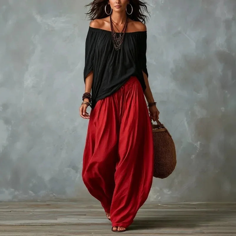 Woman wearing a black top and red pants holding a brown bag against a gray background