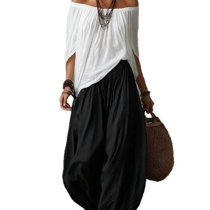 Person wearing a white off-shoulder top and black wide-leg pants, holding a brown bag on a white background.