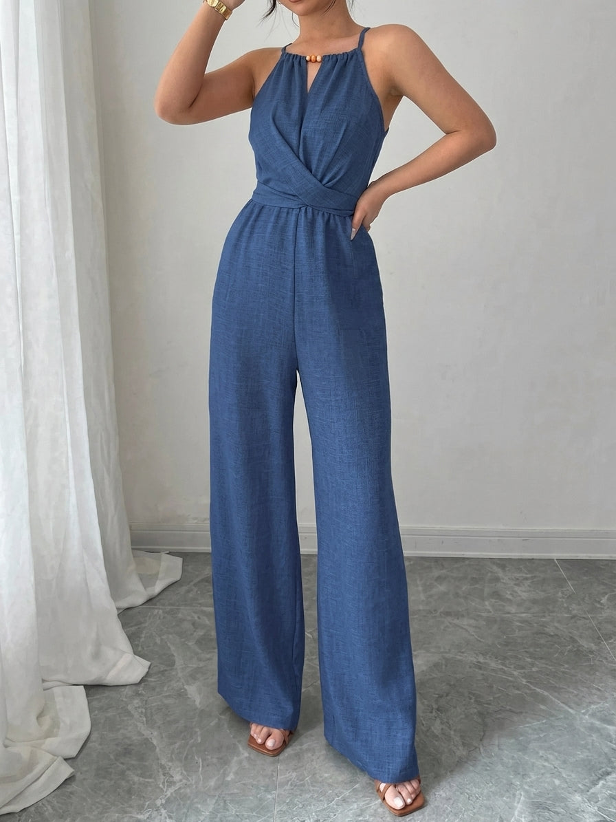 A woman modeling a blue sleeveless wide-leg jumpsuit with a halter-style neckline and twisted front detail, standing indoors against a light gray wall with a white curtain on the left.