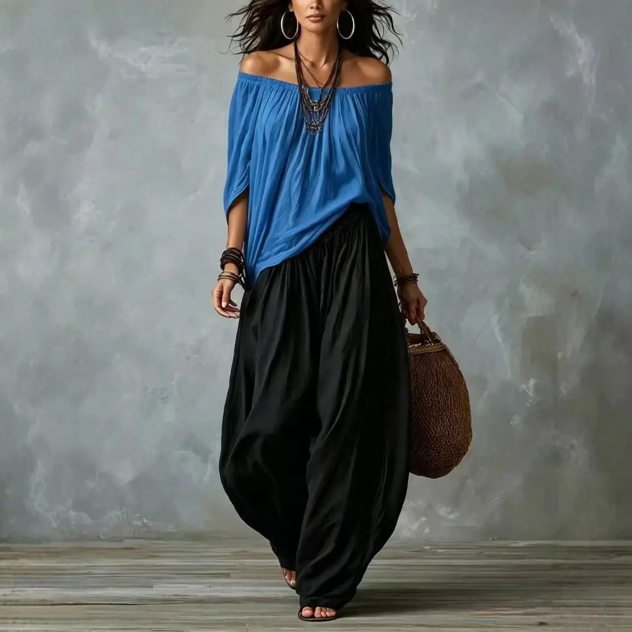 A woman modeling an off-the-shoulder blue top and loose black harem-style pants, standing against a textured gray studio wall and holding a woven brown bag.