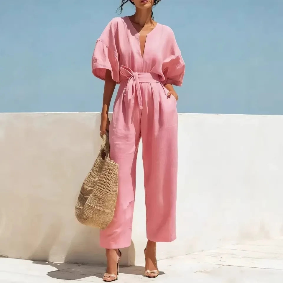 A woman models a pink linen jumpsuit with a tie waist and wide legs, holding a straw bag against a white wall and blue sky background.
