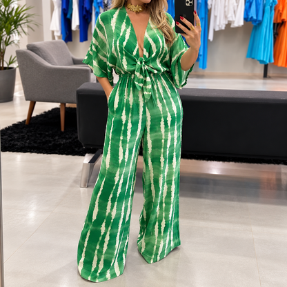 Woman showcasing an elegant green and white printed wide-leg jumpsuit featuring a plunging V-neckline, adjustable front tie, elastic waist, relaxed kimono sleeves, and practical side pockets in a stylish clothing boutique.