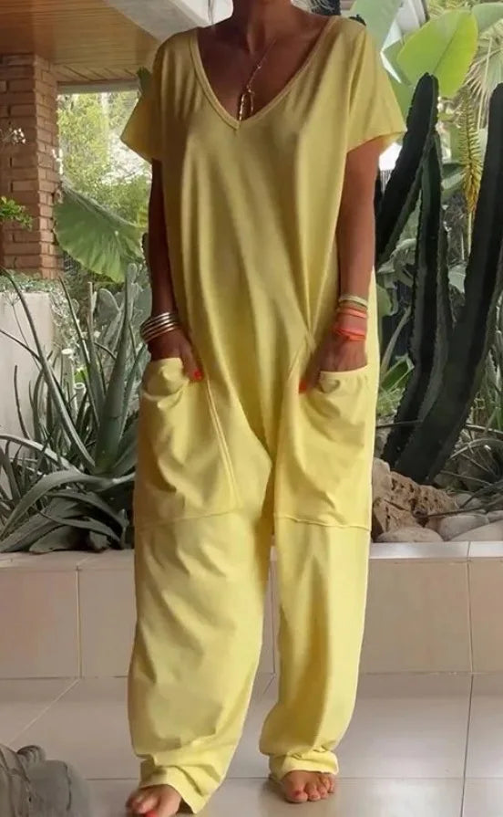 Person wearing a yellow jumpsuit standing in a garden with plants and a brick wall in the background.