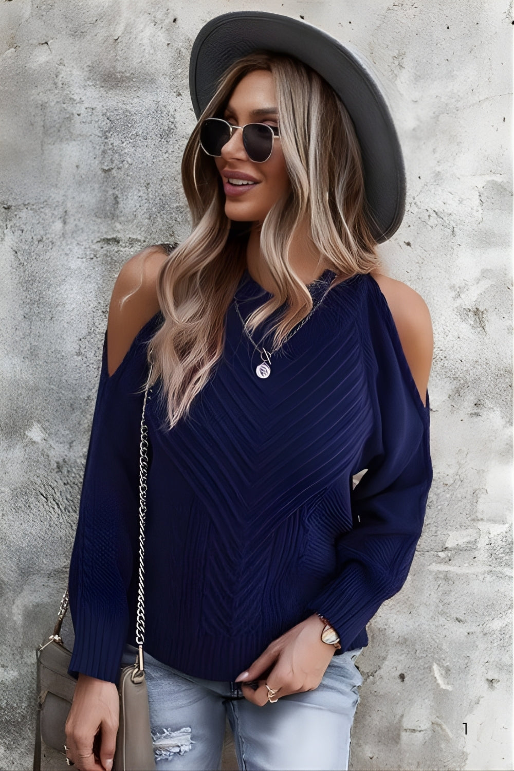 Woman wearing a navy blue off-shoulder sweater, light blue jeans, and a black hat against a textured wall.
