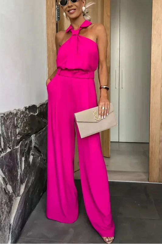 Woman in a bright pink jumpsuit holding a beige clutch in an indoor setting.