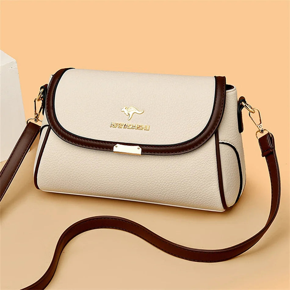 A cream-colored pebbled leather shoulder bag with dark brown trim and a gold kangaroo logo on the front flap, shown on a solid peach background with its strap draped in front.
