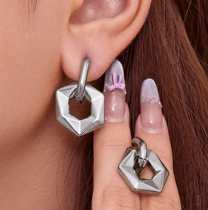 Silver geometric hoop earrings worn by a person with pink nail polish.