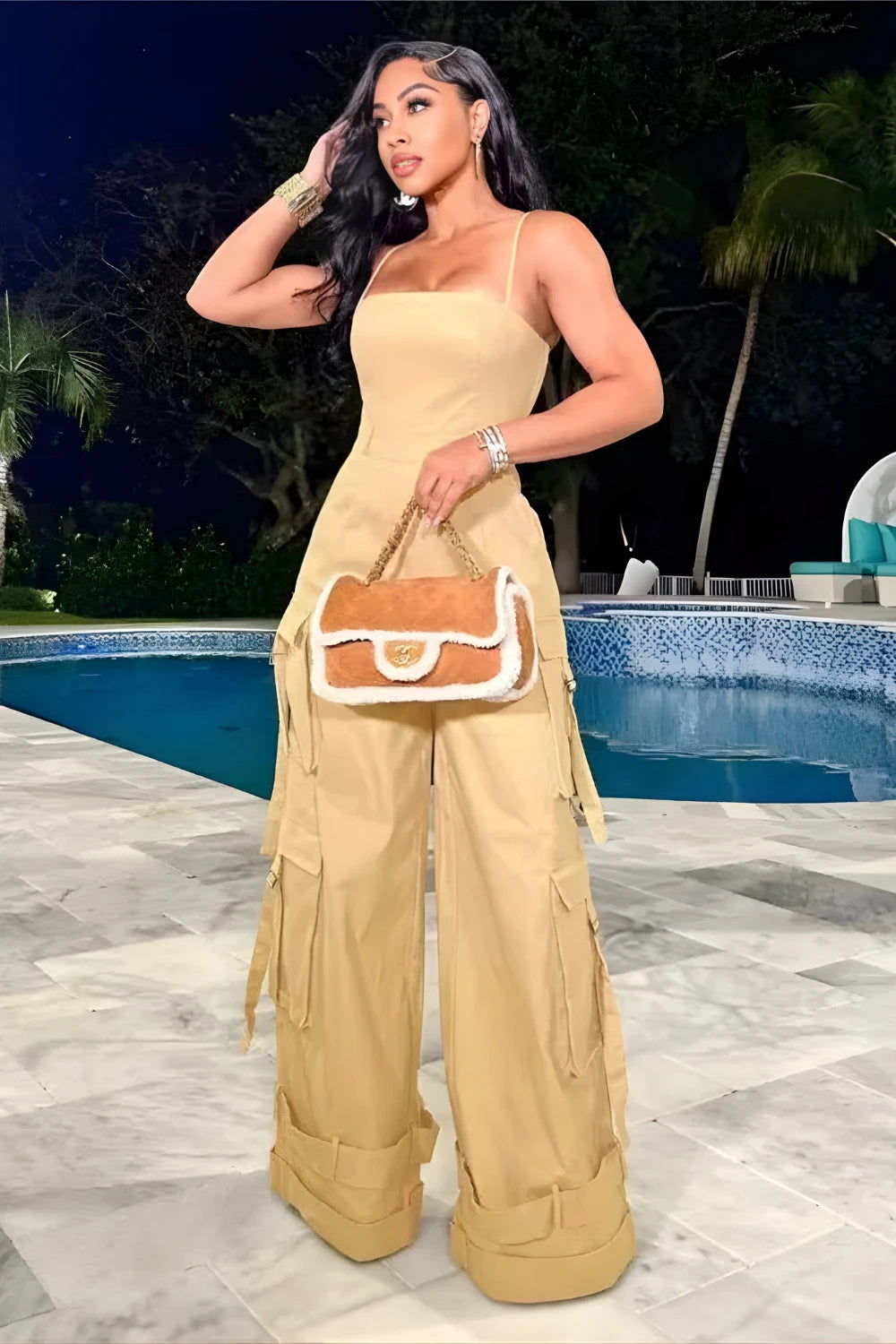 Woman in beige outfit with a brown and white handbag by a pool at night.
