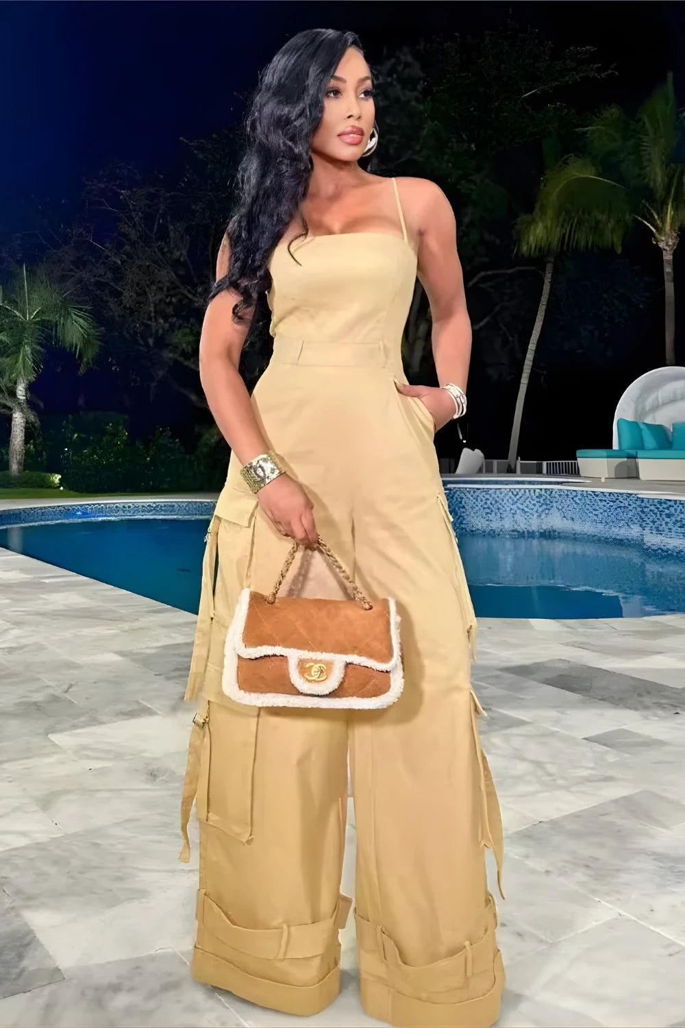 Woman in a beige outfit holding a handbag by a pool at night.