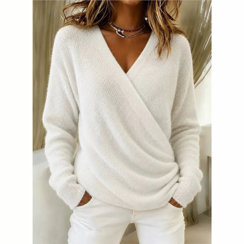 A woman wearing a fuzzy white long-sleeve V-neck wrap sweater and white pants, standing indoors with her hands in her pockets.