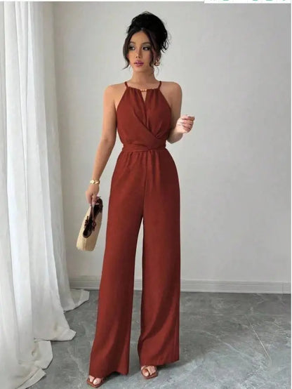 A woman models a rust-brown halter-neck jumpsuit with wide legs, holding a small beige handbag in a minimalist room with white curtains and a gray floor.