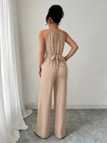 A woman viewed from behind wearing a light beige wide-leg jumpsuit with a tie-neck and tie-waist detail, standing in a room with a white curtain and gray floor.