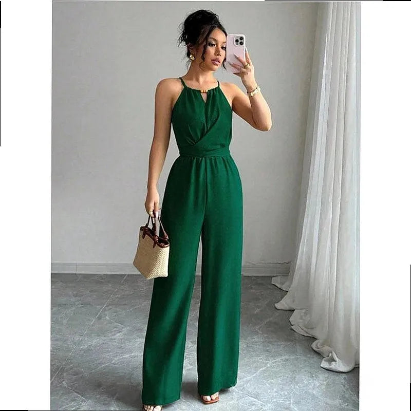 A woman takes a mirror selfie wearing a dark green halter-neck jumpsuit with a wrap-style bodice and wide legs. She is holding a small woven handbag and standing in a room with gray tiled floors and white curtains.
