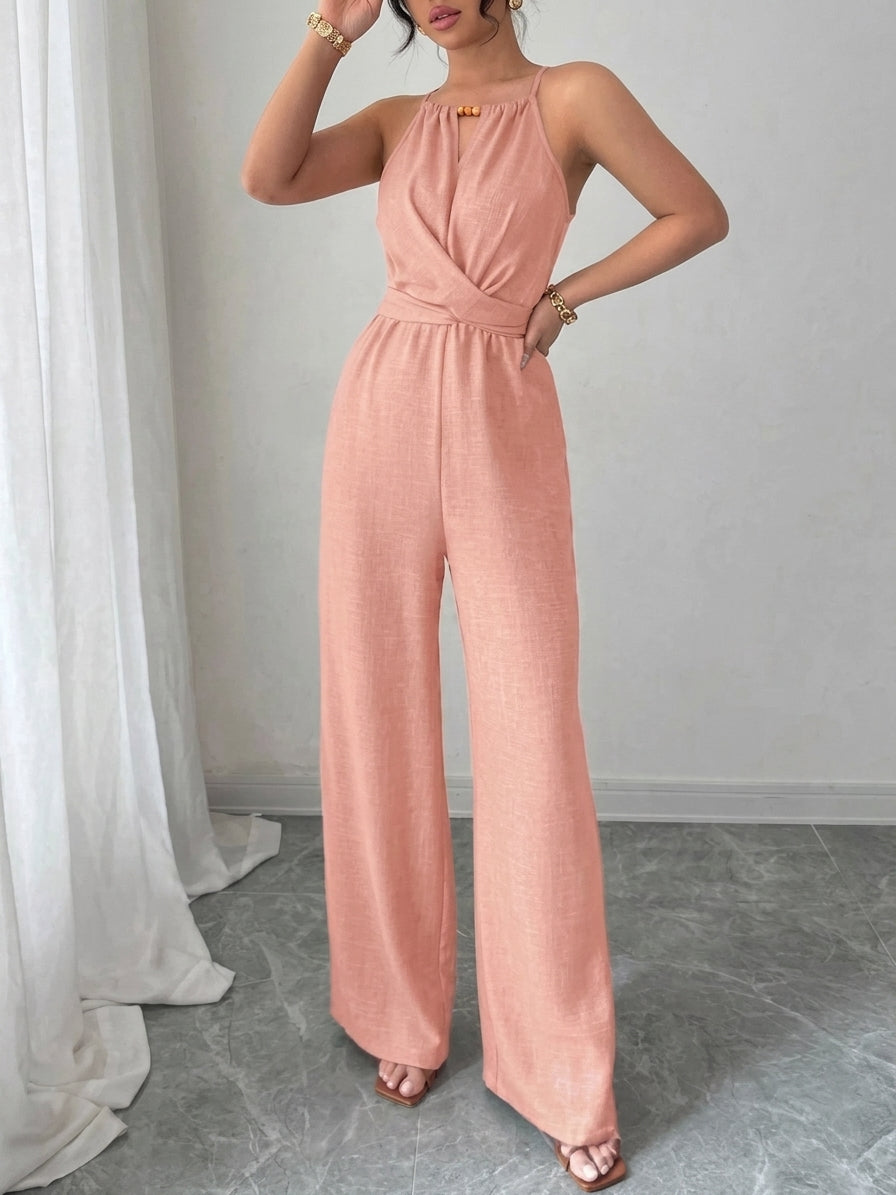 A woman models a sleeveless peach-pink wide-leg jumpsuit with a halter-style neckline and a small gold accent at the chest, standing in a room with a gray floor and white curtain.