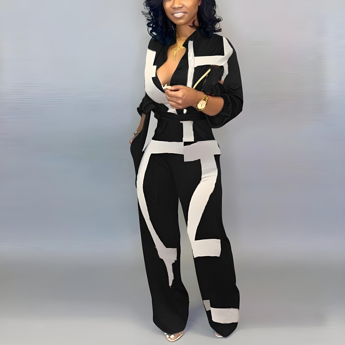 A chic black jumpsuit with striking white geometric brushstroke designs. The outfit has a belted waist, a deep V-neckline, and loose-fitting trousers. The person is accessorized with gold jewelry and shiny silver heels.