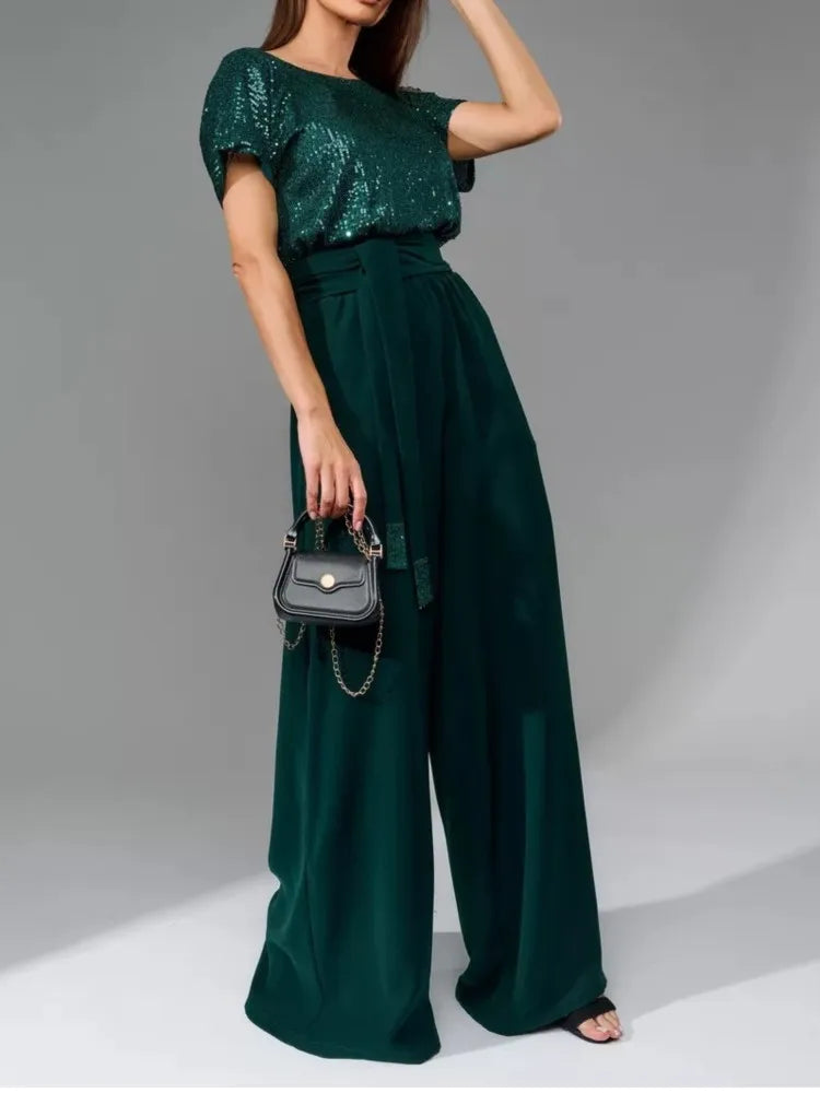 A woman models a dark green sequined short-sleeve top paired with matching wide-leg high-waisted trousers, holding a small black handbag against a gray studio background.