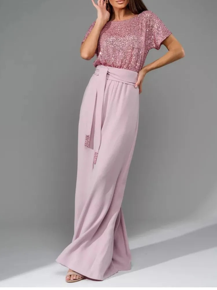 A woman modeling a two-piece outfit consisting of a short-sleeve pink sequined top and high-waisted, wide-leg dusty pink trousers with a matching tie belt, set against a plain gray studio background.