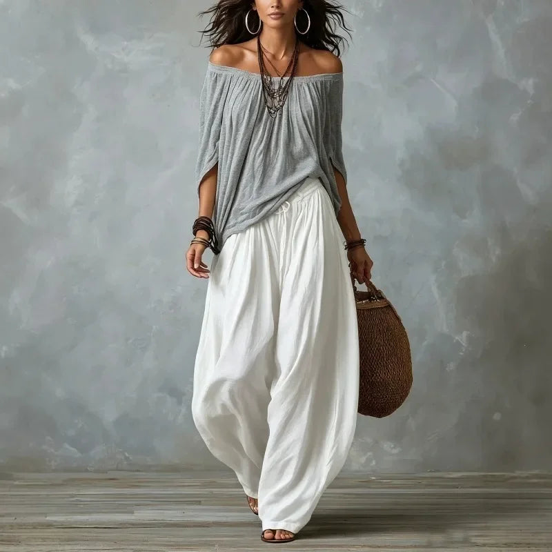 Woman wearing a gray off-shoulder top and white wide-leg pants, holding a woven bag against a gray background.