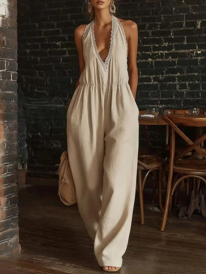 A woman models a light beige halter jumpsuit with a deep V-neckline and lace trim, set against a rustic background of a dark brick wall and wooden furniture.