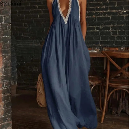A woman wearing a loose, flowing maxi dress in muted blue with white lace trim at the neckline and armholes, standing in a rustic indoor setting with a dark brick wall and wooden furniture.