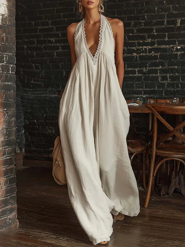 A woman models a sleeveless, off-white maxi dress with a deep V-neckline and scalloped trim, set against a rustic dark brick wall and wooden table background.