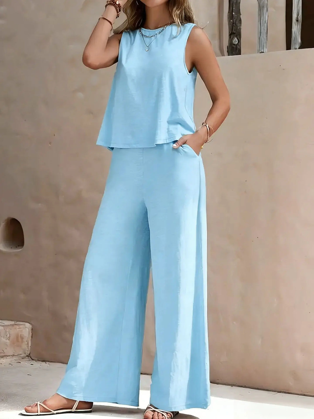 Woman wearing a light blue sleeveless top and wide-leg pants against a beige wall.