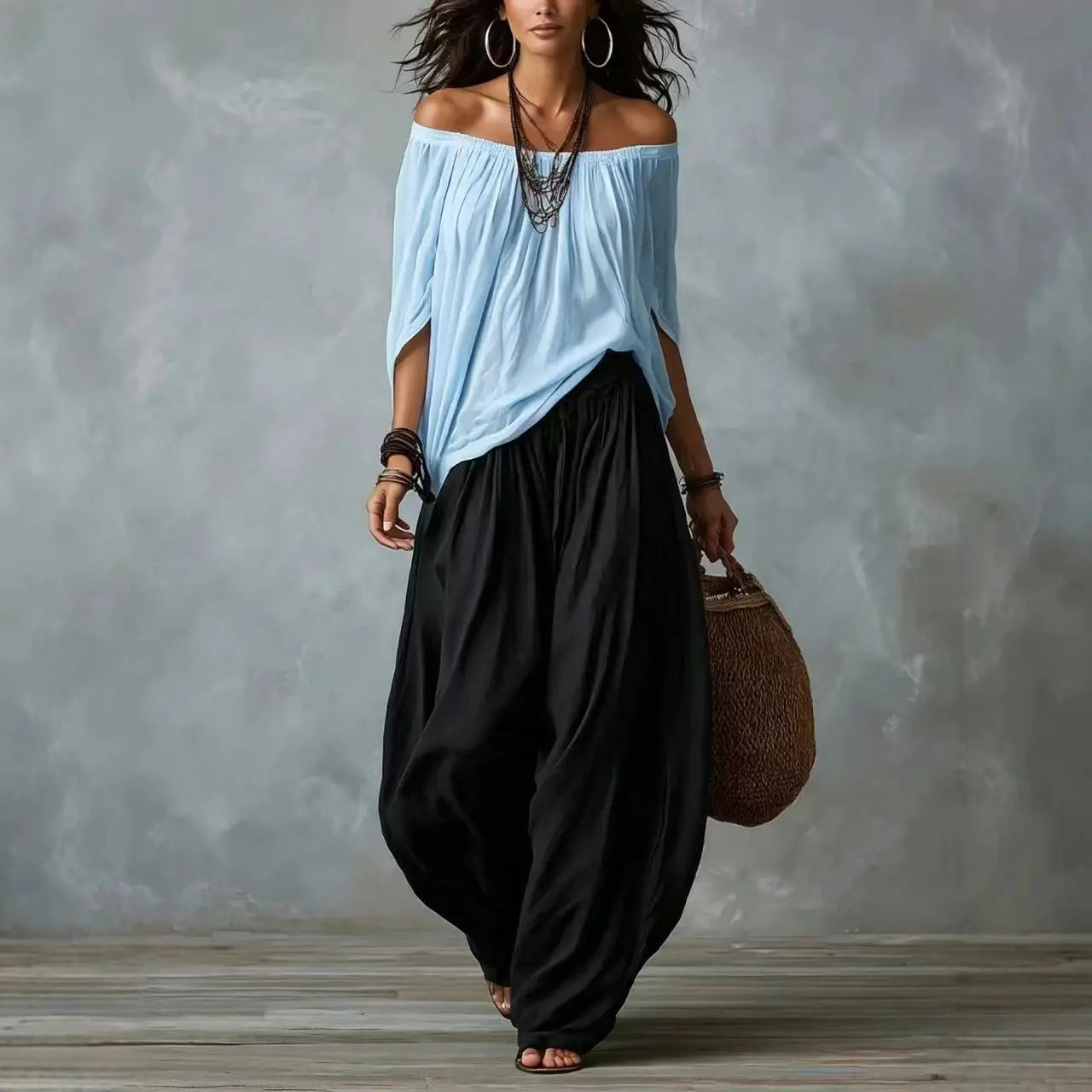 Woman wearing a light blue off-shoulder top and black wide-leg pants, holding a brown bag against a gray background.