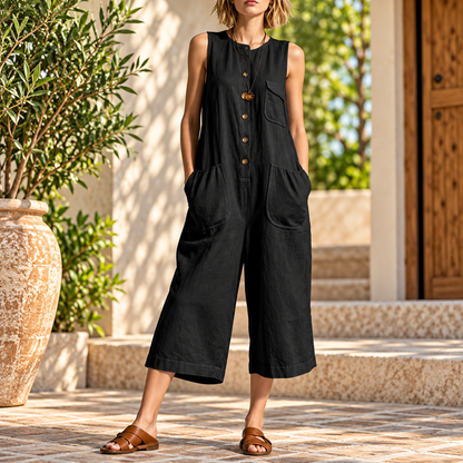 A woman wearing a black sleeveless linen-look jumpsuit with front buttons and large patch pockets, standing outdoors on stone steps beside a large potted plant.