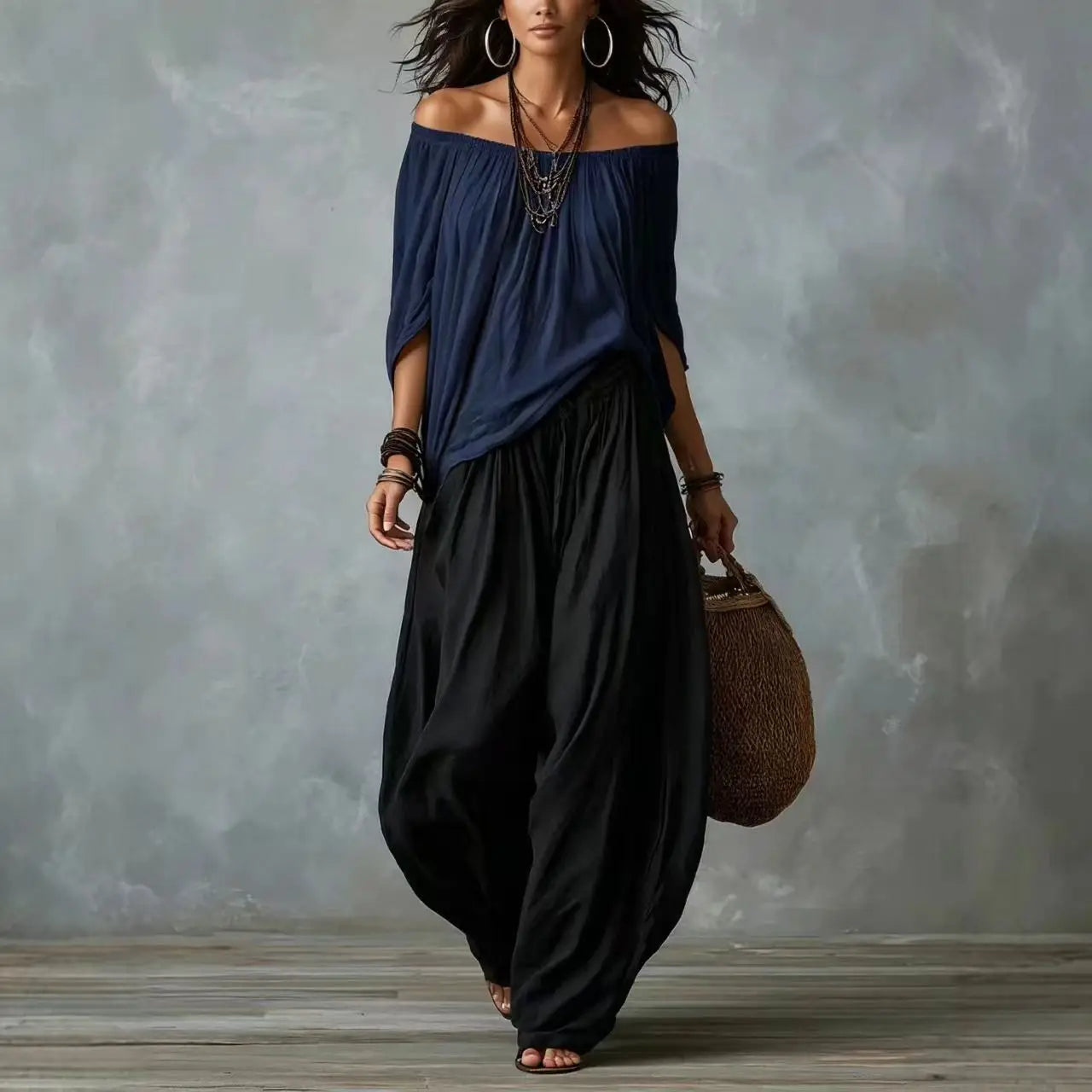 Woman wearing a navy off-shoulder top and black wide-leg pants, holding a brown woven bag against a gray textured wall.