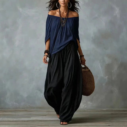 Woman wearing a navy off-shoulder top and black wide-leg pants, holding a brown woven bag against a gray textured wall.