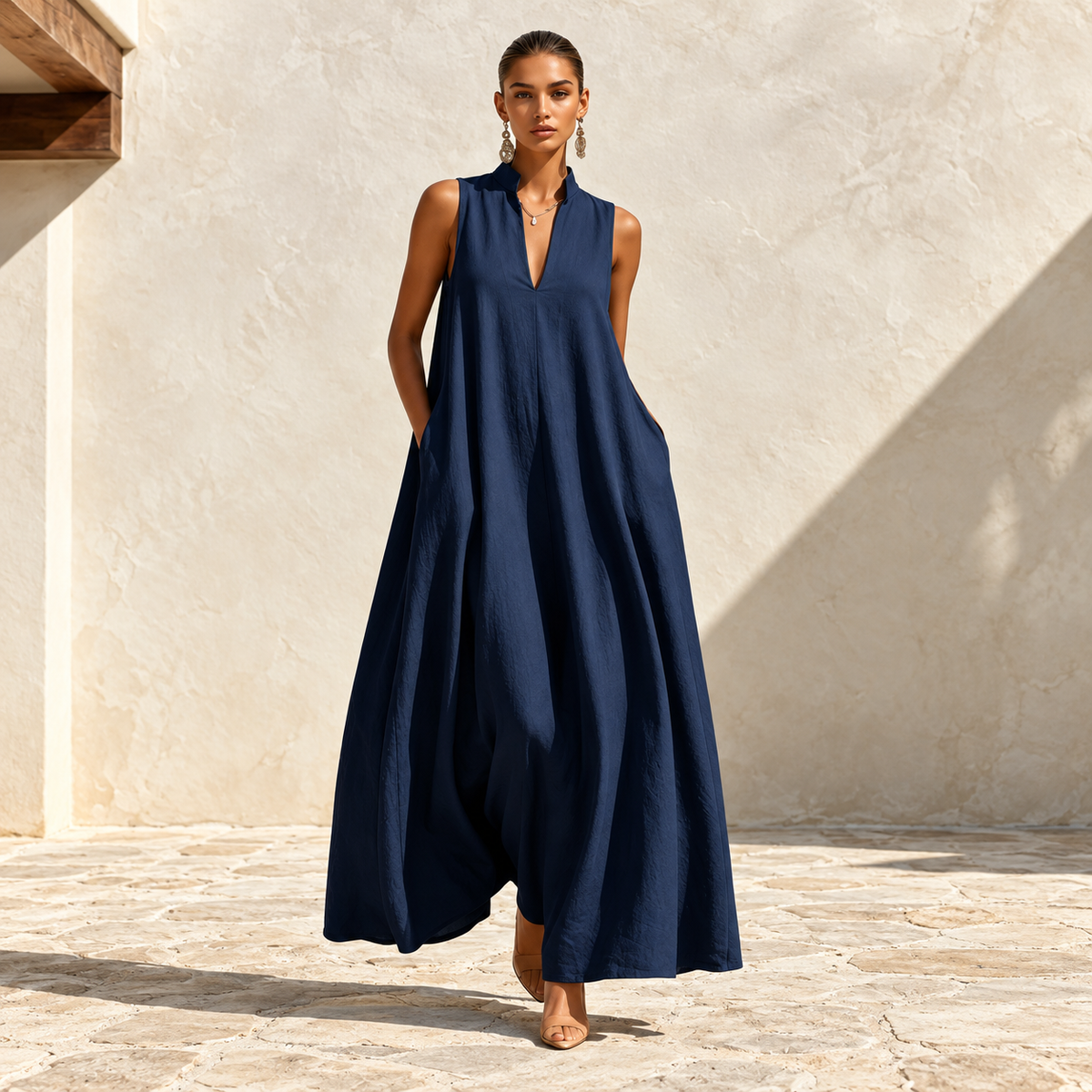 Model wearing a navy blue sleeveless maxi dress with a split V-neckline, side pockets and a flowing silhouette in a sunlit ivory courtyard.