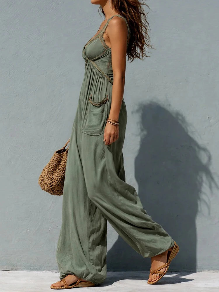 A woman wearing a sage green wide-leg jumpsuit with thin straps and a gathered waist, walking against a textured light blue-gray wall. She is carrying a woven straw bag and wearing tan flat sandals.