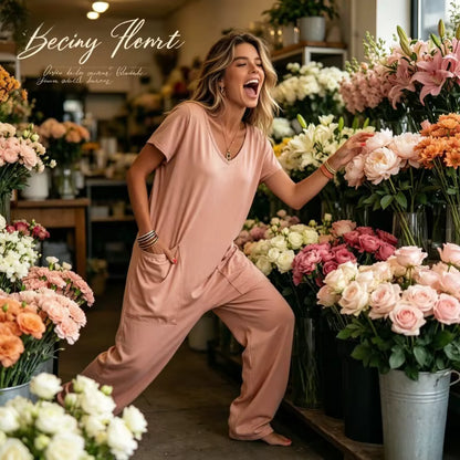 A woman modeling a dusty rose short-sleeve lounge set in a flower shop, surrounded by colorful bouquets of roses and lilies. The brand name 'Beciny Flomt' is visible in the top left corner.