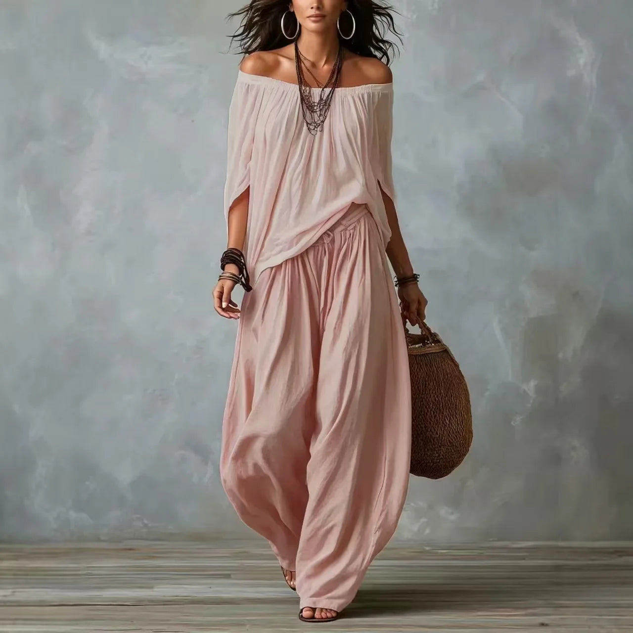 Woman wearing a flowing pink dress holding a woven bag against a textured gray wall.