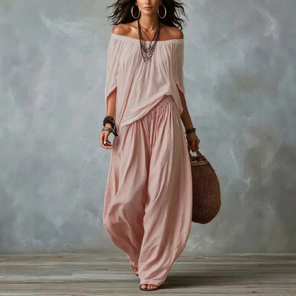 Woman wearing a flowing pink dress holding a woven bag against a textured gray wall.