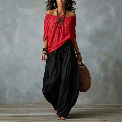 Woman wearing a red off-shoulder top and black wide-leg pants, holding a brown bag against a gray textured wall.