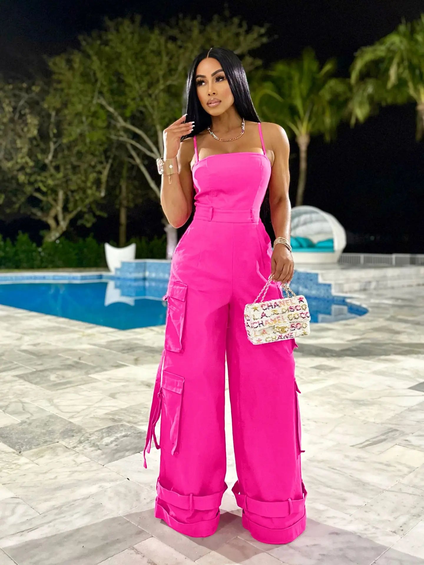Woman in a bright pink jumpsuit holding a Chanel handbag by a pool at night.