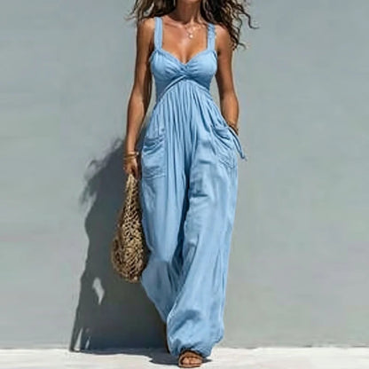 A woman wearing a light blue sleeveless wide-leg jumpsuit with a gathered bust and side pockets, standing against a pale gray wall and holding a woven straw bag.