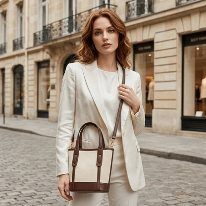A woman stands on a cobblestone street wearing a cream-colored blazer and trousers, carrying a structured tote bag with cream fabric and dark brown leather trim and handles.