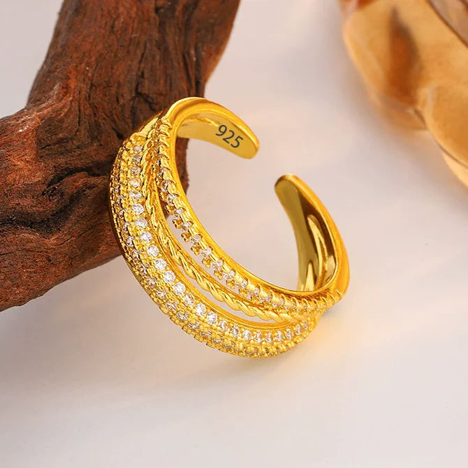 A gold-toned open cuff ring with three parallel rows of small clear stones, photographed on a white surface with a piece of brown wood prop. The inside of the ring is stamped with '925'.
