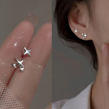 Silver star-shaped earrings shown on a finger and worn on an ear against a neutral background.