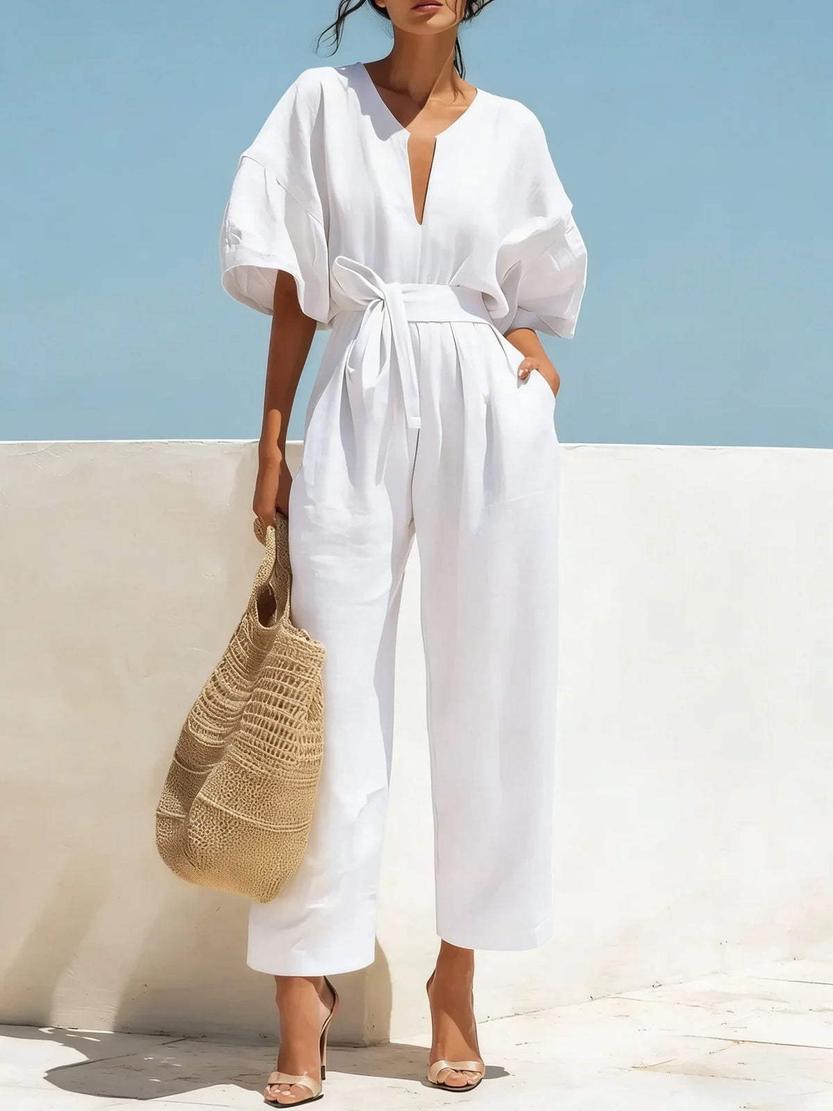 Woman wearing a white jumpsuit holding a straw bag against a blue sky.